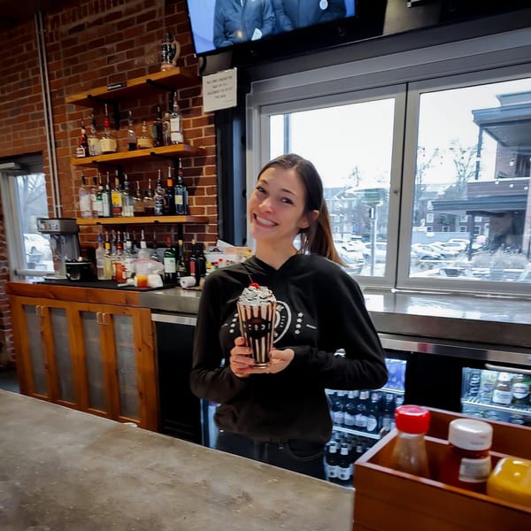 A smiling Hobo's employee holds a chocolate milkshake with whipped cream and a cherry at the bar.