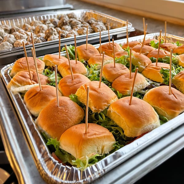 Hobo's catering tray of mini sliders with toothpicks and a tray of meatballs in the background.