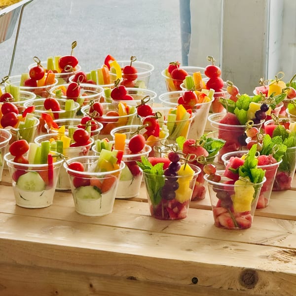 Rows of plastic cups filled with fresh fruit and vegetable appetizers on a wooden table.
