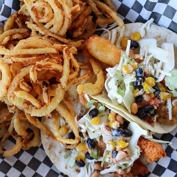 Two Hobo's fish tacos with corn, beans, and cabbage, served with a side of crispy onion rings.