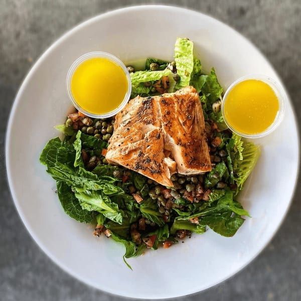 A Hobo's salmon salad with greens, capers, and two cups of dressing in a white bowl.