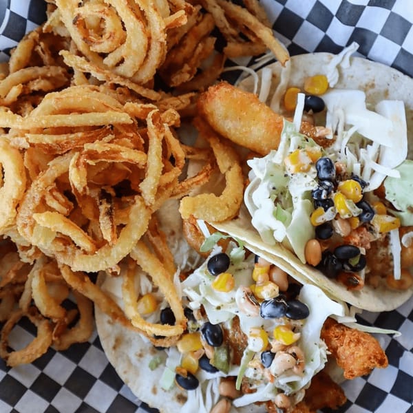 Two fish tacos with corn, beans, and cabbage, alongside a pile of crispy onion rings.