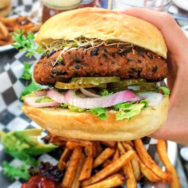 A hand holds a Hobo's black bean burger with lettuce, red onion, pickles, and sprouts, with fries below.
